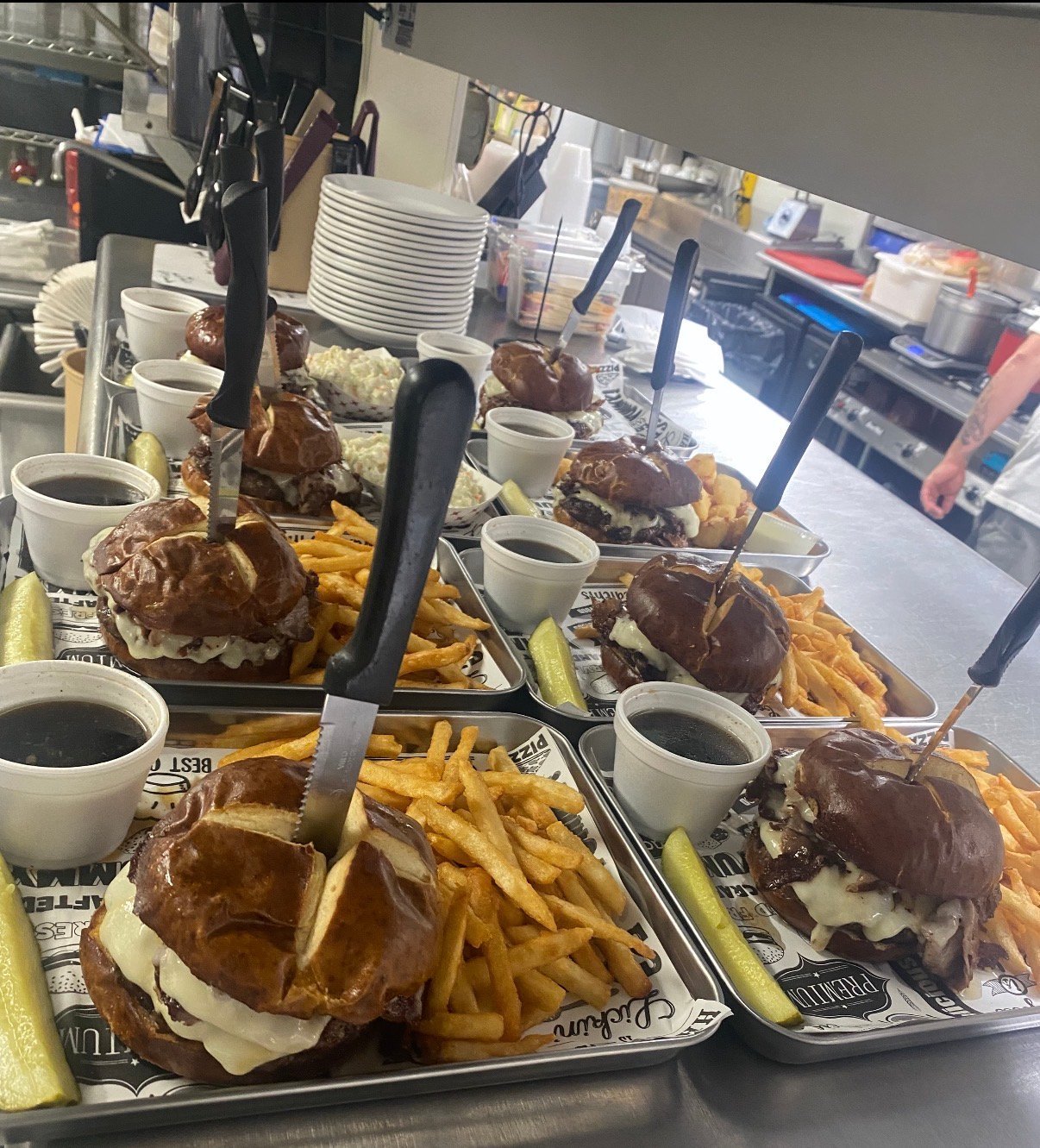 Multiple burger and fries meal trays displayed in a restaurant counter window with stacked plates and beverages visible