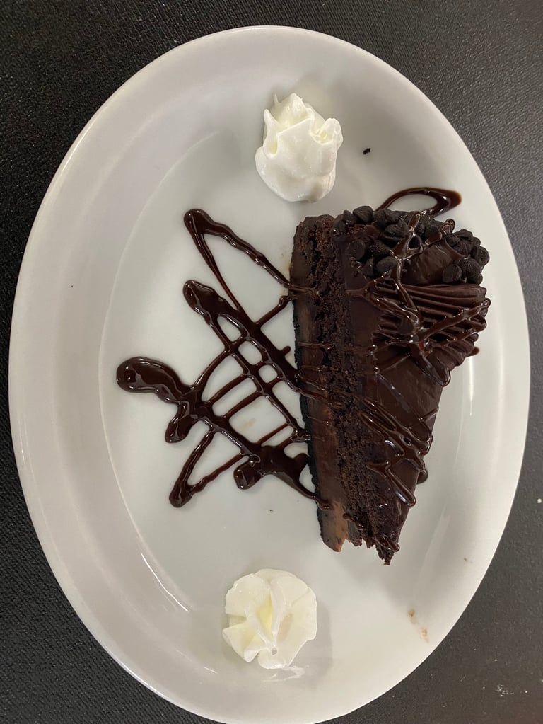 Slice of rich chocolate cake with chocolate drizzle on white plate, topped with whipped cream dollops