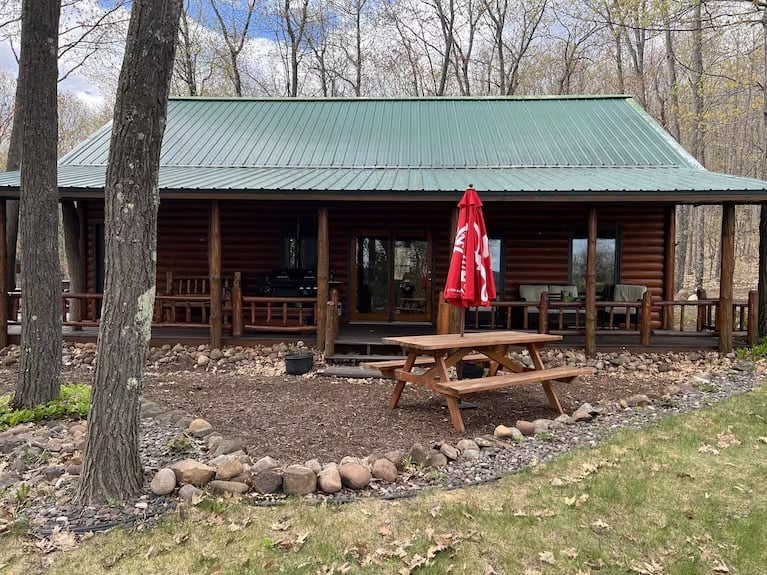 Rustic log cabin with green metal roof, front porch, and picnic table surrounded by bare trees in a wooded setting