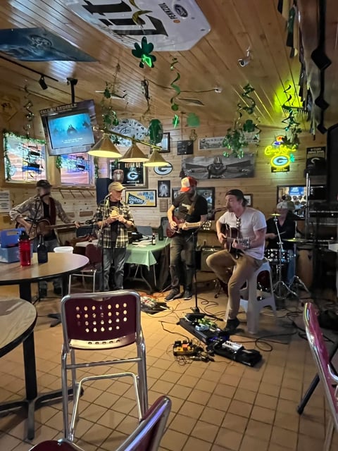 Band performing in a casual venue with wooden walls, hanging plants, and neon signs, with audience members seated nearby