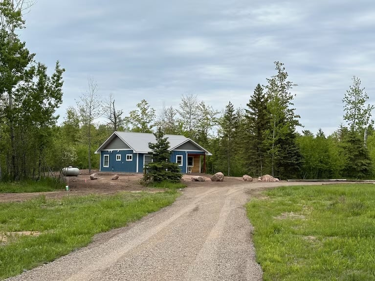 Blue cottage-style house with gray roof situated on rural gravel driveway surrounded by green fields and evergreen trees