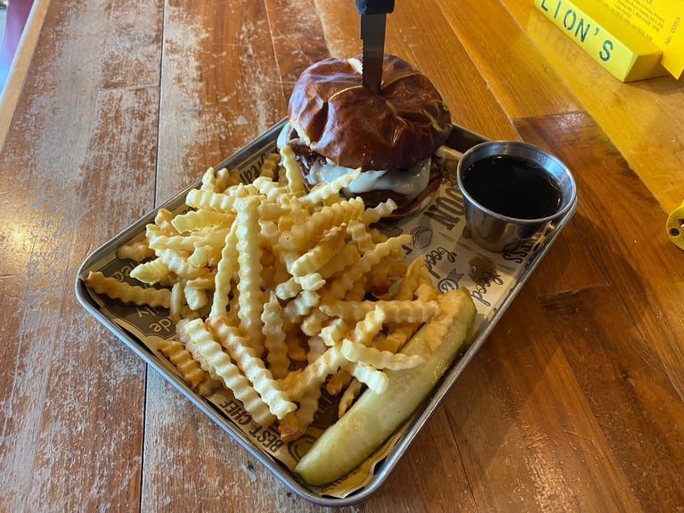 A metal tray holding a burger with toppings and wavy-cut french fries, served with a small cup of black dipping sauce on a wooden table