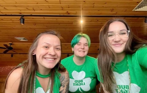 Three young women wearing green and white clothing with shamrock designs, smiling at camera in wooden interior