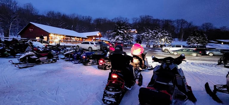 Snowmobiles gathered at dusk in snowy parking lot near illuminated lodge with bare trees in background