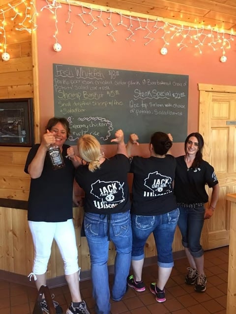 Four people wearing matching black Jacks Wisconsin shirts pose together in front of a menu board in a wooden restaurant interior with holiday lights