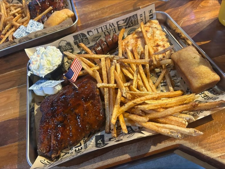 Metal tray of barbecued ribs with golden french fries, coleslaw, and cornbread on a wooden table