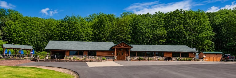Wooden barn complex with multiple buildings and stone foundation surrounded by green fields and dense forest under blue sky