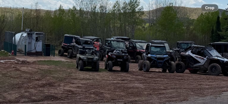 Row of black and gray recreational vehicles parked in dirt lot with trees and buildings in background
